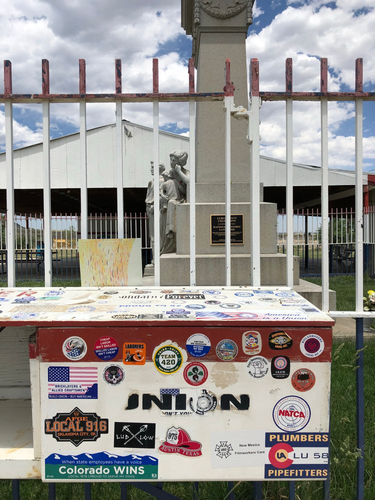 Photograph of the side of a box(?) outside the fence surrounding the Ludlow Monument, which is covered with various union stickers both general and for local unions. June 2018.