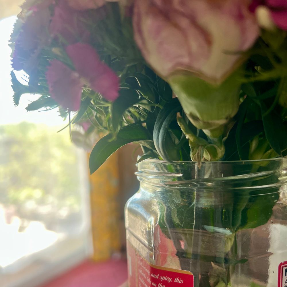 A tall, octagonal glass jar holds a bunch of pink flowers. In its previous life it was a jar containing pasta sauce. I wonder if it is happier now?