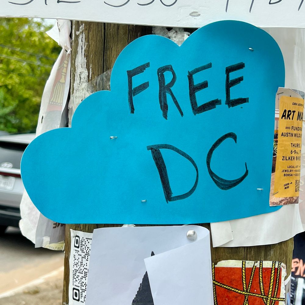 Photo of a telephone pole covered with paper flyers, the only one of which is fully visible is a hand-drawn sign on blue poster board that’s been cut into a cloud-ish shape. FREE DC is written across it in black sharpie. Photo taken August 31, 2025 in Austin, Texas. 