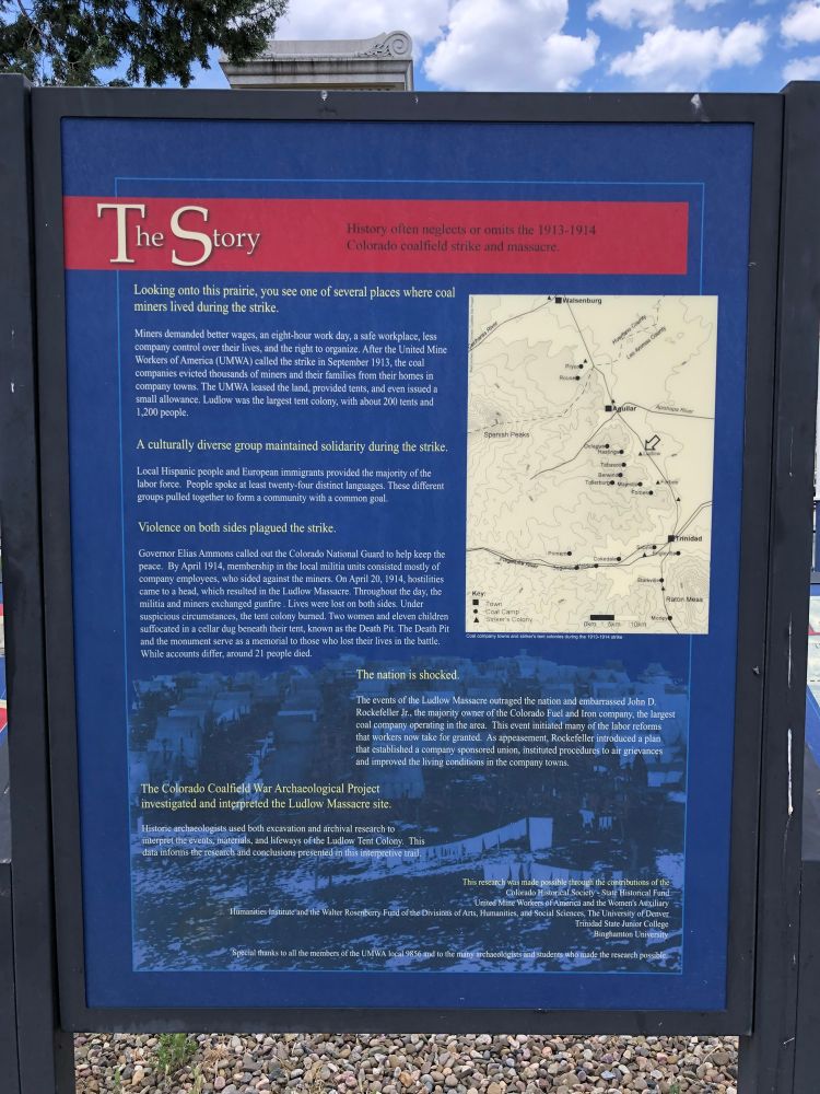 June 2018 photograph of an interpretive/information sign at the Ludlow Memorial. Text (truncated): The Story History often neglects or omits the 1913-1914 Colorado coalfield strike and massacre. Looking onto this prairie, you see one of several places where coal miners lived during the strike. Miners demanded better wages, an eight-hour work day, a safe workplace, less company control over their lives, and the right to organize. After the United Mine Workers of America (UMWA) called the strike in September 1913, the coal companies evicted thousands of miners and their families from their homes in company towns. The UMWA leased the land, provided tents, and even issued a small allowance. Ludlow was the largest tent colony, with about 200 tents and 1,200 people. A culturally diverse group maintained solidarity during the strike. Local Hispanic people and European immigrants provided the majority of the labor force. People spoke at least twenty-four distinct languages. These different groups pulled together to form a community with a common goal. Violence on both sides plagued the strike. Governor Elias Ammons called out the Colorado National Guard to help keep the peace. By April 1914, membership in the local militia units consisted mostly of company employees, who sided against the miners. On April 20, 1914, hostilities came to a head, which resulted in the Ludlow Massacre. Throughout the day, the militia and miners exchanged gunfire . Lives were lost on both sides. Under suspicious circumstances, the tent colony burned. Two women and eleven children suffocated in a cellar dug beneath their tent, known as the Death Pit. The Death Pit and the monument serve as a memorial to those who lost their lives in the battle. While accounts differ, around 21 people died.