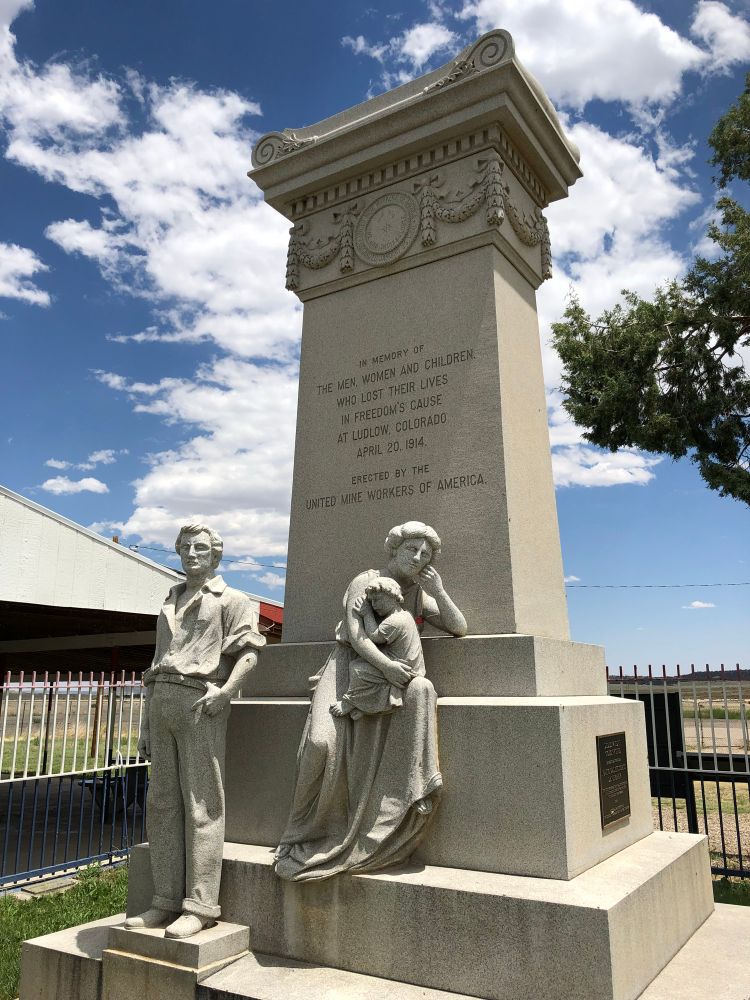 Photo from June 2018. The Ludlow Memorial in Trinidad, Colorado (Las Animas County), the site of The Ludlow Massacre where striking miners and some of their families were killed. The monument is a tall die, base, and cap style with an inscription on one if the broad sides. Below the inscription are figures of a man standing slightly apart from a woman holding a young child on her lap. 