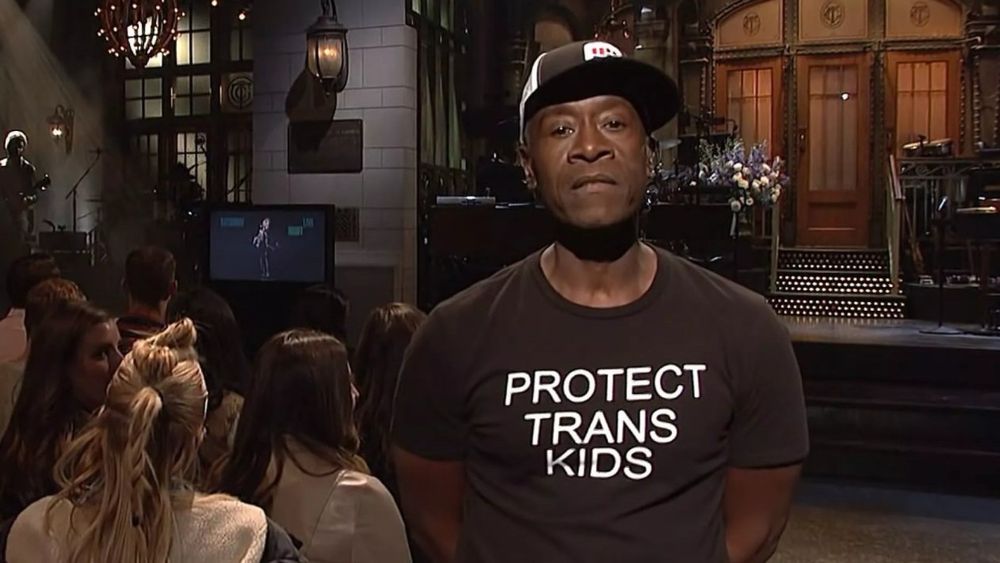Don Cheadle appearing on Saturday Night Live in 2019. He is about to introduce the musical guest. The stage is behind him, audience members behind him have their backs to him. He is looking into the camera. He’s wearing a baseball cap of some kind and a dark shirt with the phrase PROTECT TRANS KIDS in white letters across his chest.