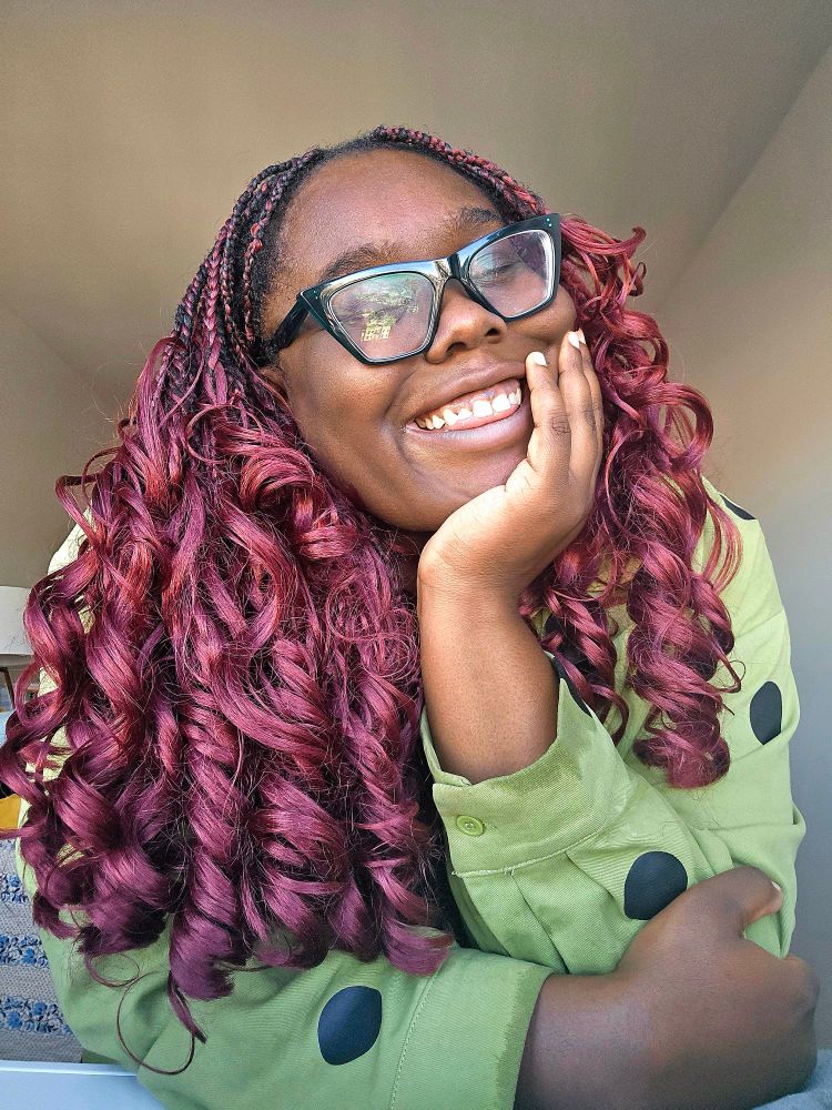 Dark skin Black woman with red wine curly box braids, green trapezoid glasses, and a green shirt with black polka dots smiles into the camera with her hand on her chin.