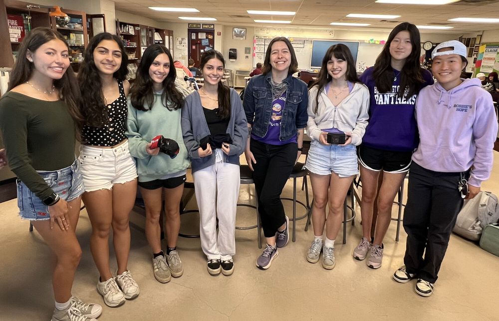 Seven high schoolers, one teacher, all smiles. The young women are holding their prototype