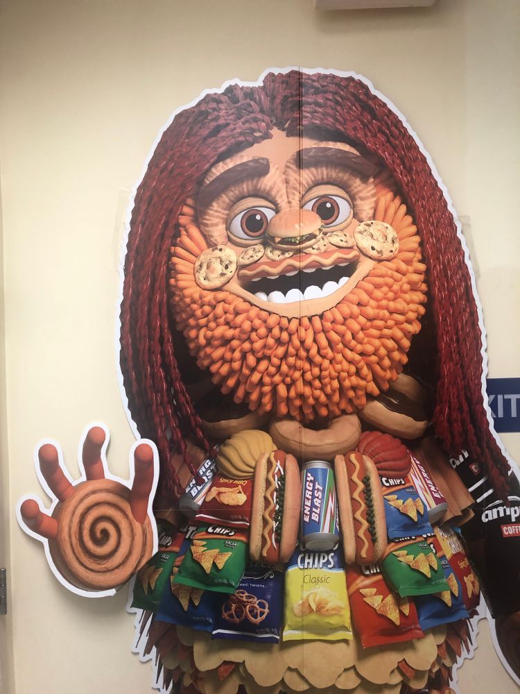 A terrifying monstrosity cardboard cutout standup, made of collaged animated depictions of snack food. His face is a pufferfish of hot dogs, leering with a chocolate chip cookie smile, and he reaches for you with one cinnamon roll hand, grasping with his hot dog fingers.