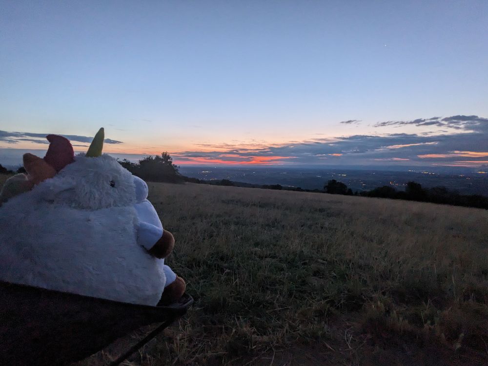 Photo de paysage pris depuis le haut d'un petit mont, ciel très matinal avec les lueurs du soleil derrière les nuages a l'horizon. Au premier plein a gauche, une énorme peluche de licorne assise sur une petite chaise de camping regarde le paysage