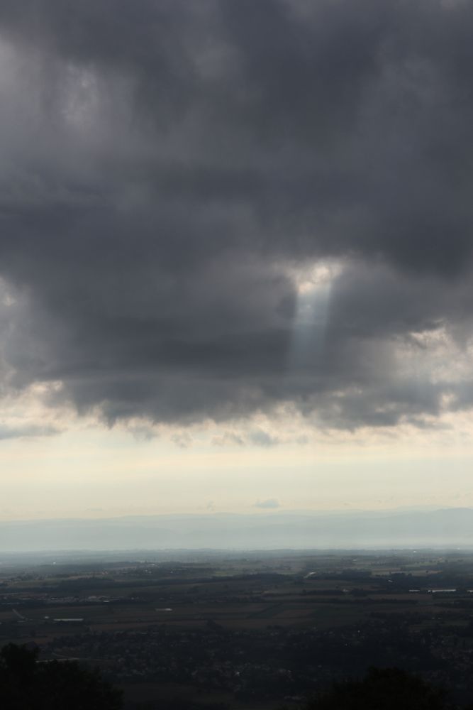 Photo verticale d'un paysage, prise du haut d'un petit mont. Une vaste plaine occupe le premier quart en bas. L'horizon est clair et forme une bande blanche sur le second quart. Au dessus, un ciel très sombre, avec une trouée qui laisse passer un timide mais épais rayon de soleil