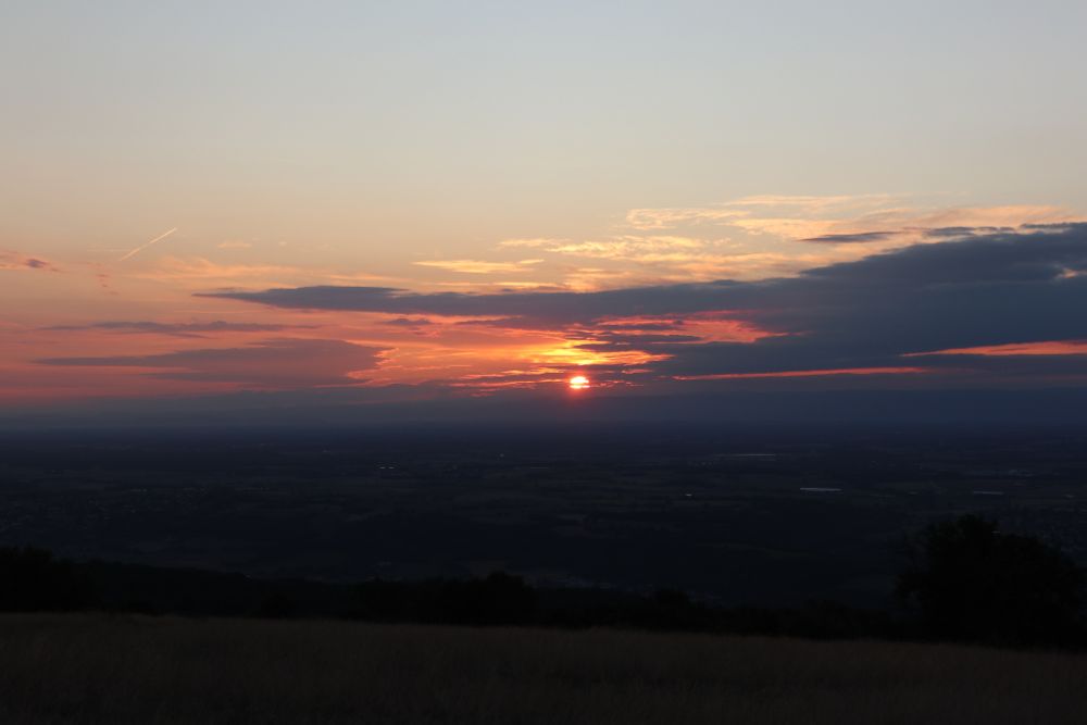 Plan large d'un paysage à l'aube. La moitié basse de l'image est une vaste plaine dans la pénombre. À l'horizon le soleil pointe le nez à travers les nuages, le ciel crée une bande bien rose et orange. Au dessus, le ciel est dégagé et clair, qui occupe le dernier quart de l'image