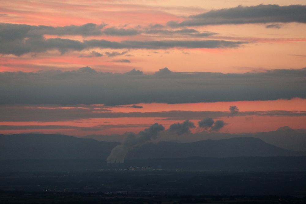 Photo d'un paysage à l'aube. La moitié basse de la photo est occupée par une plaine dans la pénombre. Au fond, on devine la centrale nucléaire du Bugey et ses 4 énormes cheminées qui émettent un imposant panache, poussé par le vent vers la droite de l'image. 
Derrière, les formes de grosses collines se découpent sur un clien rose pale parsemé de nuages qui s'étirent en longueur