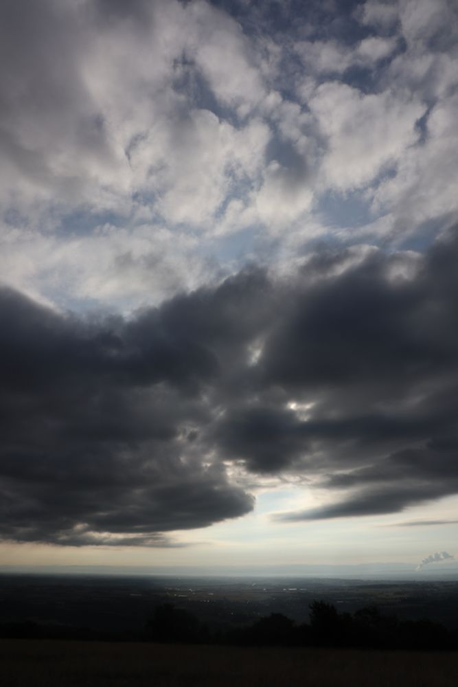 Photo verticale de paysage. En bas, une vaste plaine plongée dans l'obscurité. Au dessus, d'épais nuages très sombres qui masquent le soleil. Il y a quand même une grosse percée à travers les nuages et une partie de la plaine apparait très éclairée en contraste du reste très sombre.