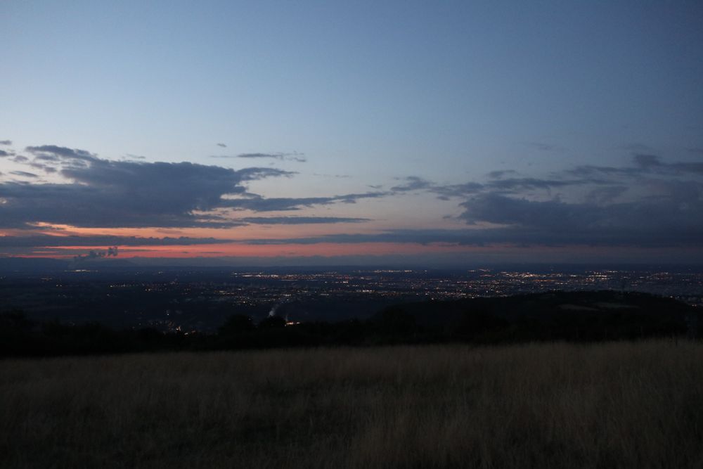 Photo paysage pris du haut d'un petit mont, à l'aube (6h du matin).Au premier plan, une praire d'herbes sèches. La terre est encore dans la pénombre, on voit les lumières de Lyon plus loin, l'horizon qui rosit avec quelques épais nuages foncés, et un ciel dégagé et clair au dessus