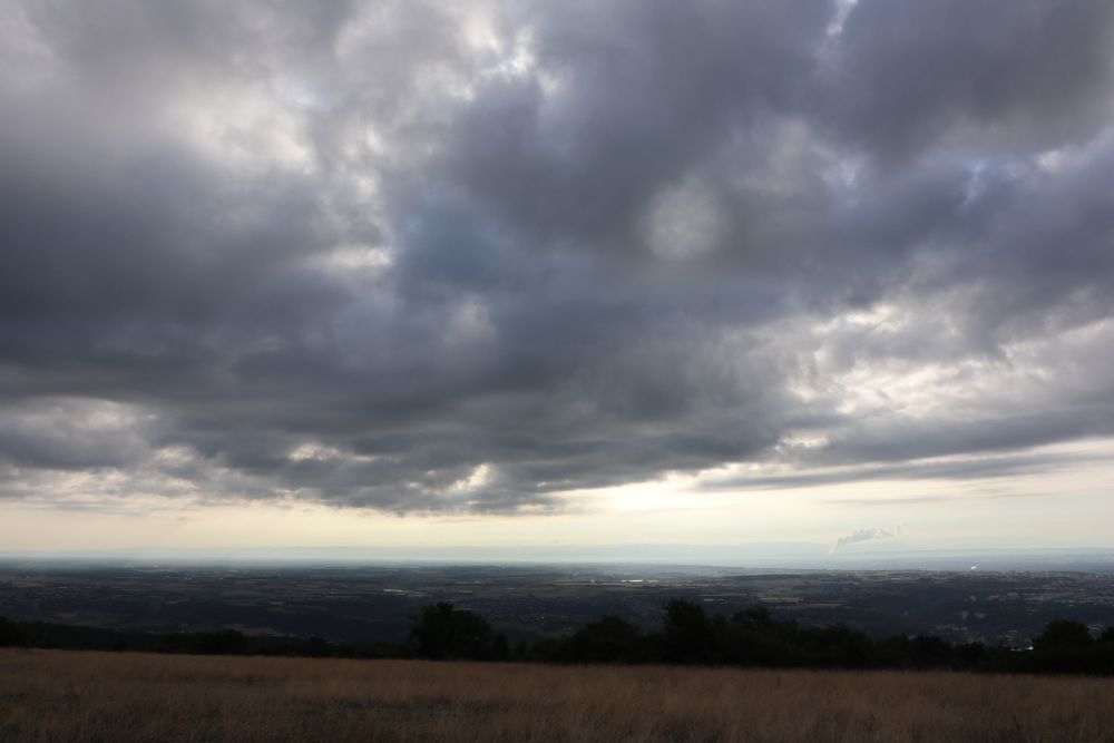 Photo paysage au petit matin (8h20), au premier plan un pré d'herbe sèches qui descend vers une barrière d'arbre. Très loin derrière (et plus bas) une vaste pleine. Au dessus, immense, un ciel très nuageux et écrasant occupe la plus grand partie de l'image