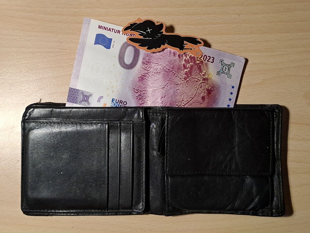 An empty, black wallet with a purple 0€ bill sticking out of it and a sticker of a dead crow on top.