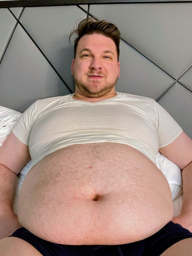 A large man lays in bed with his head propped up on the headboard facing the camera. A tight white shirt is too small and only covers his chest. 