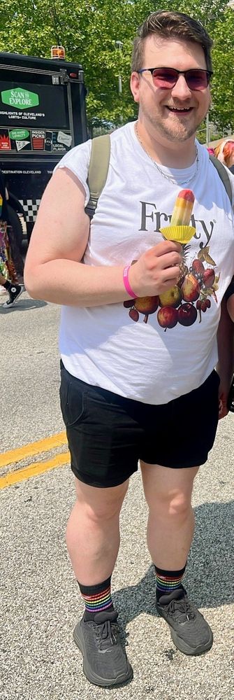 Large man holding a popsicle wesring black shorts and a sleeveless white graphic shirt that has fruit and the word "fruity on it