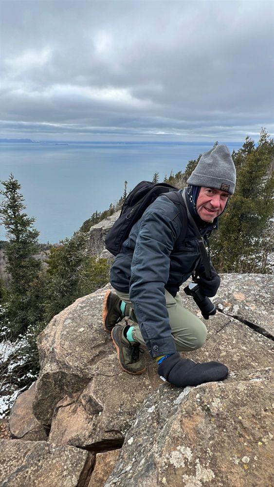 The poster, warmly dressed in jacket, mitts, & toque, and wearing a backpack, climbs over granite rocks. Far below, and extending to the horizon, is Lake Superior.