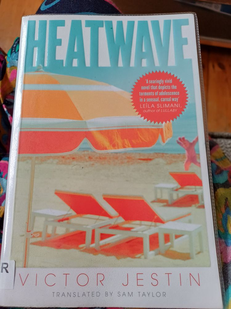 Cover of book, 'Heatwave'in blue at top. A scene of a beach, two deckchairs,  a beach umbrella in various shades of orange, and a pink life-size bunny rabbit.