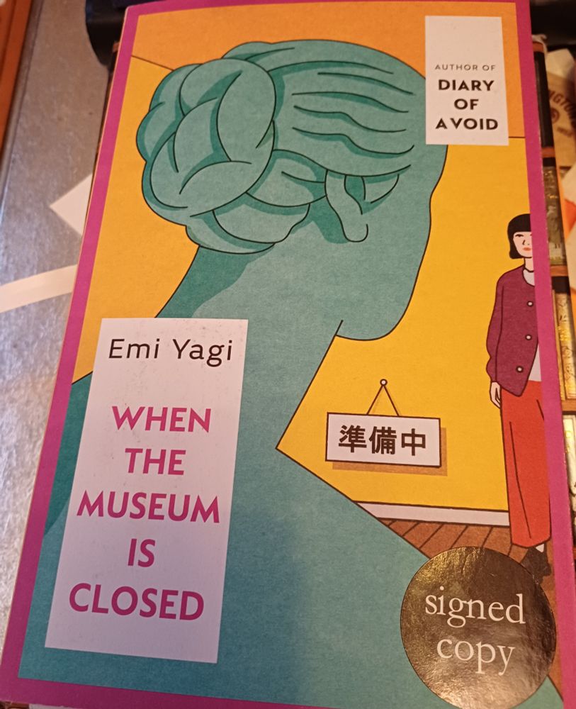 Book cover. Title of the book is in pink writing 'When The Museum Is Closed' in an enclosed box with white background in lower left of book along with author's name Emi Yagi in black. Cover has the back of a blue statue of Venus looking towards a young Japanese girl, who is wearing a loose red jacket and red trousers and looking back at the statue . There is a sign hanging on the yellow wall written in Japanese