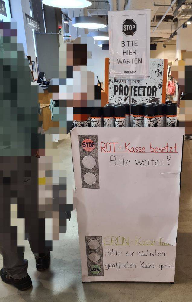 Kassenbereich eines Kaufhauses, dort wird das Ampelsystem der Kassen erklärt.

Oben ein kleines ausgedrucktes Schild:
"STOP
BITTE HIER WARTEN"

Darunter ein großes handgeschriebenes Schild:
"ROT = Kasse besetzt
Bitte warten!
GRÜN = Kasse frei
Bitte zur nachsten geöffneten Kasse gehen"