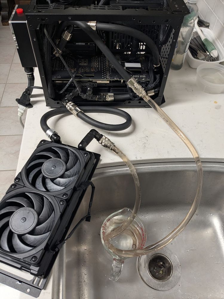 a small liquid-cooled PC next to a kitchen sink.  one radiator and fan assembly is removed and sitting on the counter.  two clear hoses are attached to the PC internal black hoses, and are running into a water-filled measuring cup in the sink.