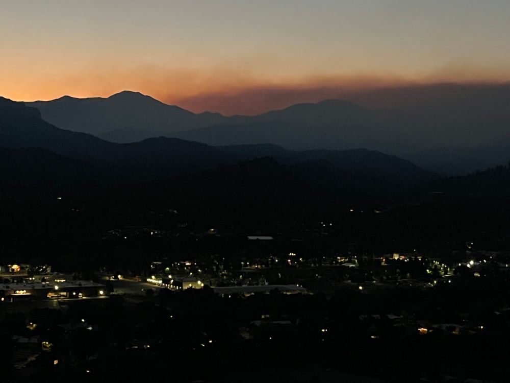 In the foreground is a small town lit up at night. In the background are mountain ranges with dark colored smoke pouring over one of the mountains, covering up the view downwind.