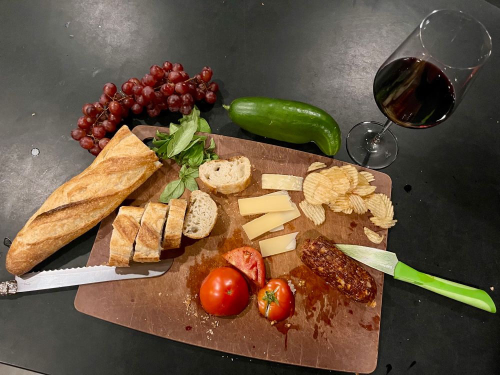 Brown rectangular cutting board with a baguette cut up in the slices Parmesan cheese slices a fresh tomato, fresh basil, chorizo, sausage, potato chips, cucumber, and red grapes. On the side of the cutting board is a bread, knife and a sharper knife for the tomato and chorizo as well as a glass of red wine.