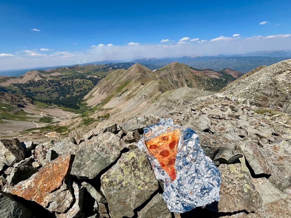 Slice of pepperoni pizza on tinfoil sitting on a pile of rocks on a summit of a mountain. The horizon is noticeably slanted.
