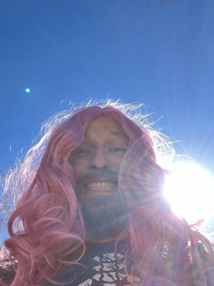 A very stupid looking beardo with long pink hair. Brilliant blue sky in background with sun blasting over the shoulder.