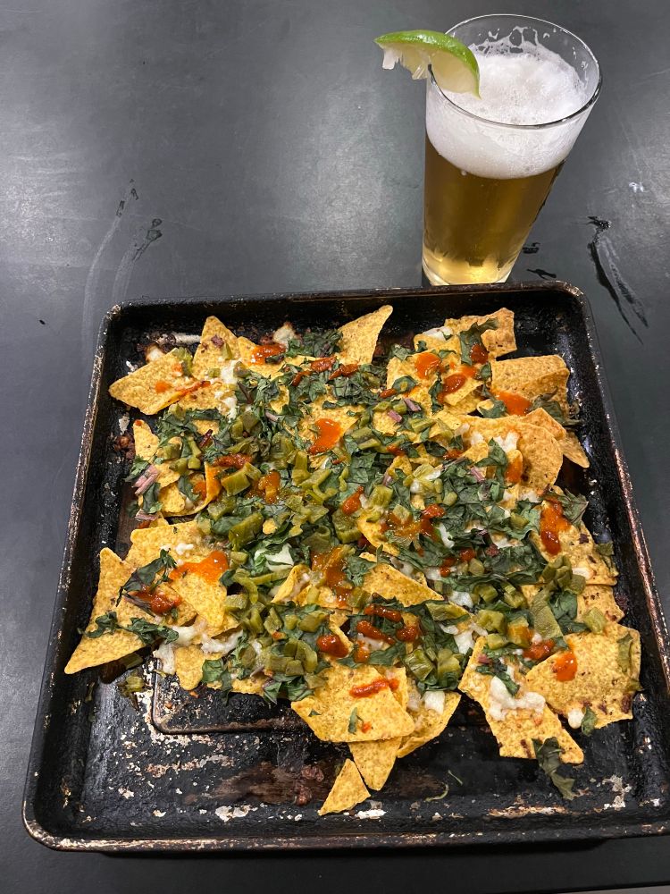 A black cooking pan covered in nachos consisting of yellow corn chips, melted queso, chard fresh from the garden, green chilis, salsa. A clear pint glass holding a Mexican logger and a slice of lime.