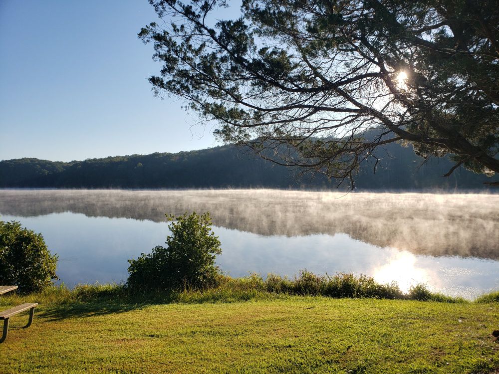 Green trees and low hills surround a misty lake reflecting the morning sun
