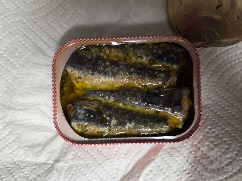 A close up shot of the open can of sardines in butter. Four small fish are arranged in an alternating pattern in the golden liquid, flecks of parsley and bits of garlic coat them. The can is resting on a paper towel.