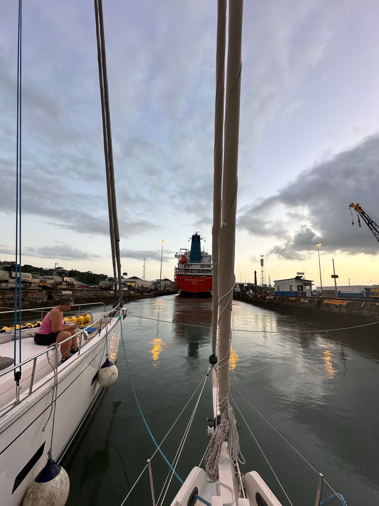 Two rafted sailboats point at the transom of a red container ship called Barrington Island. The waters in the lock are calm. The sunset and overhead lights reflect on the water. This was our Panama Canal transit. 