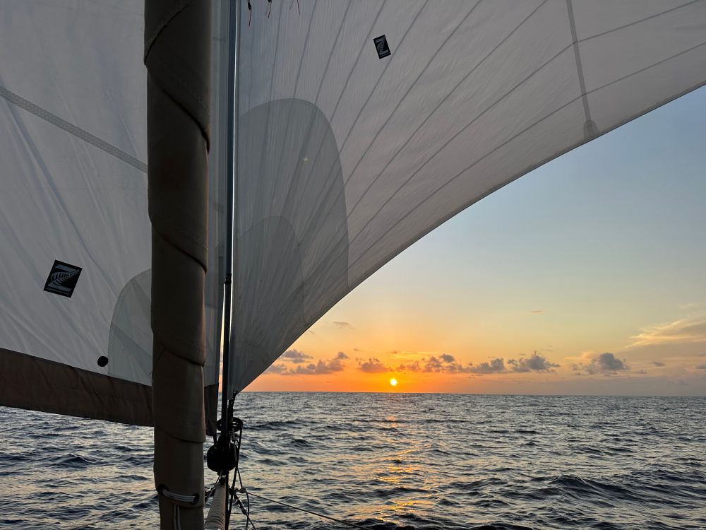 A glowing ocean sunset peeks out from under a poled-out light air sail. The jib is flying to port. Calm seas with enough wind to sail. It’s glorious. 