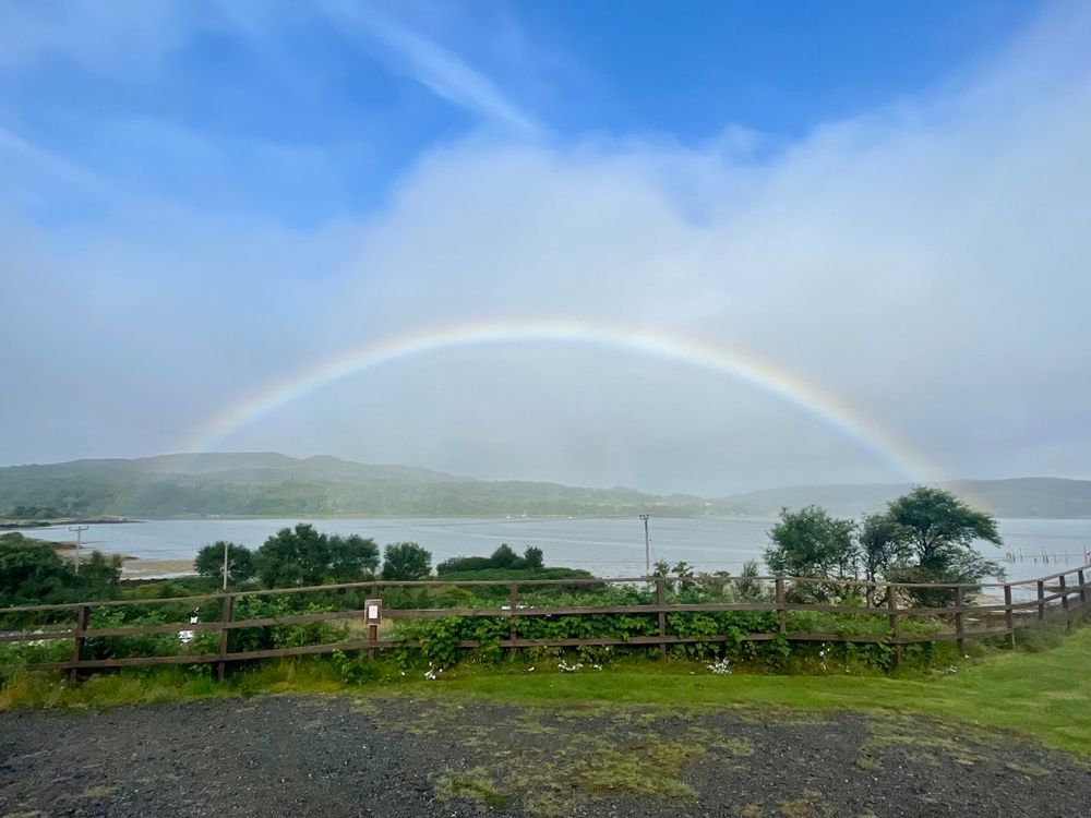 Een regenboog boven een baai, de lucht is blauw met een paar wolkenflarden. In de voorgrond staan wat bomen en planten en een houten hek. In de achtergrond zijn beboste heuvels 