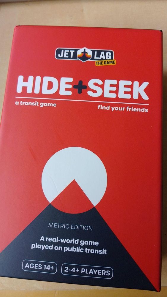Doos van het spel Hide+Seek van Jet Lag:The Game. De doos is rood en heeft de tekst "A transit game, find your friends. A real-world game played on public transit"