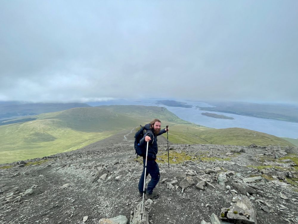 Ik (een 35 jarige man met blond haar en een baard) sta op berg. Ik heb een rugzak om en houd twee wandelstokken in de lucht terwijl ik naar de camera kijk. In de achtergrond is een berglandschap te zien met een zee-inham waar verschillende eilanden in liggen