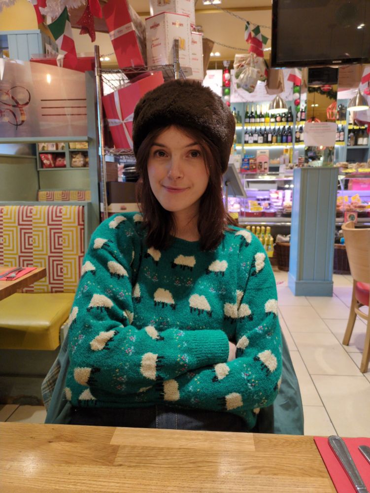 The skeeter's girlfriend in an Italian deli, wearing a green wool jumper with little sheep on it and a faux fur headband