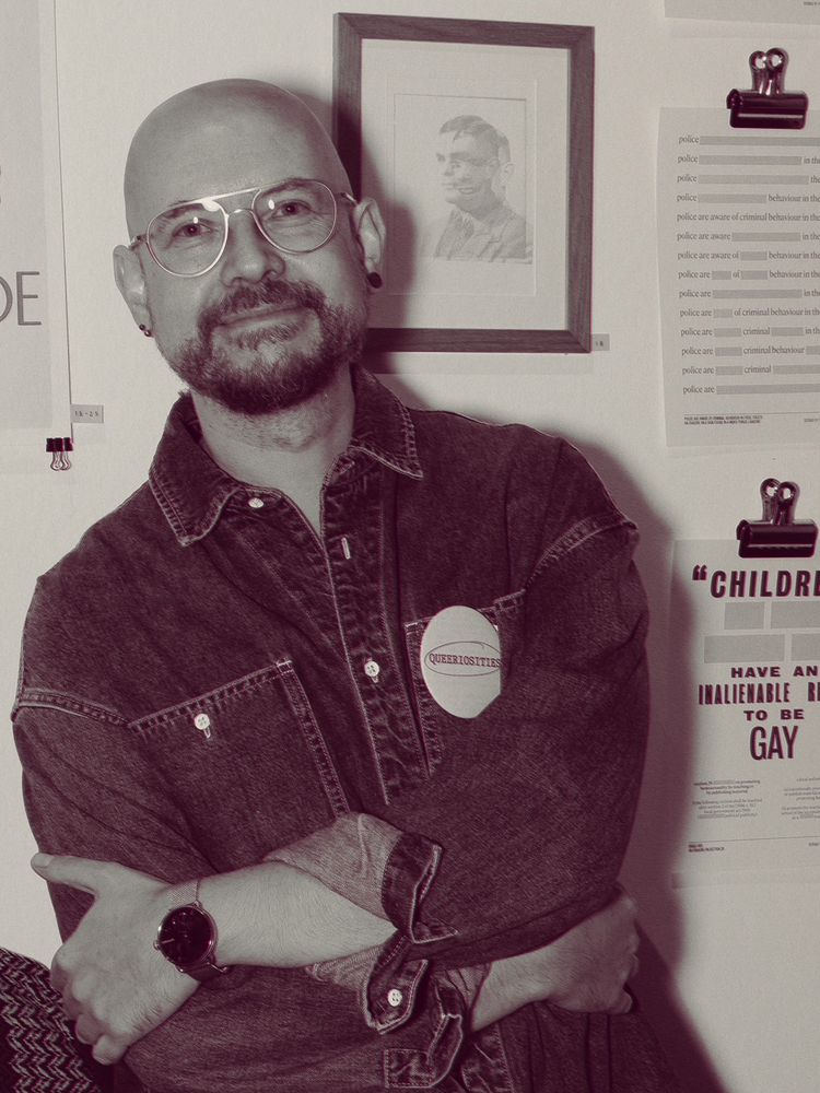 A photo of Peter Collins, arms folded and smiling to camera. He’s standing in front of a wall displaying his artwork. 