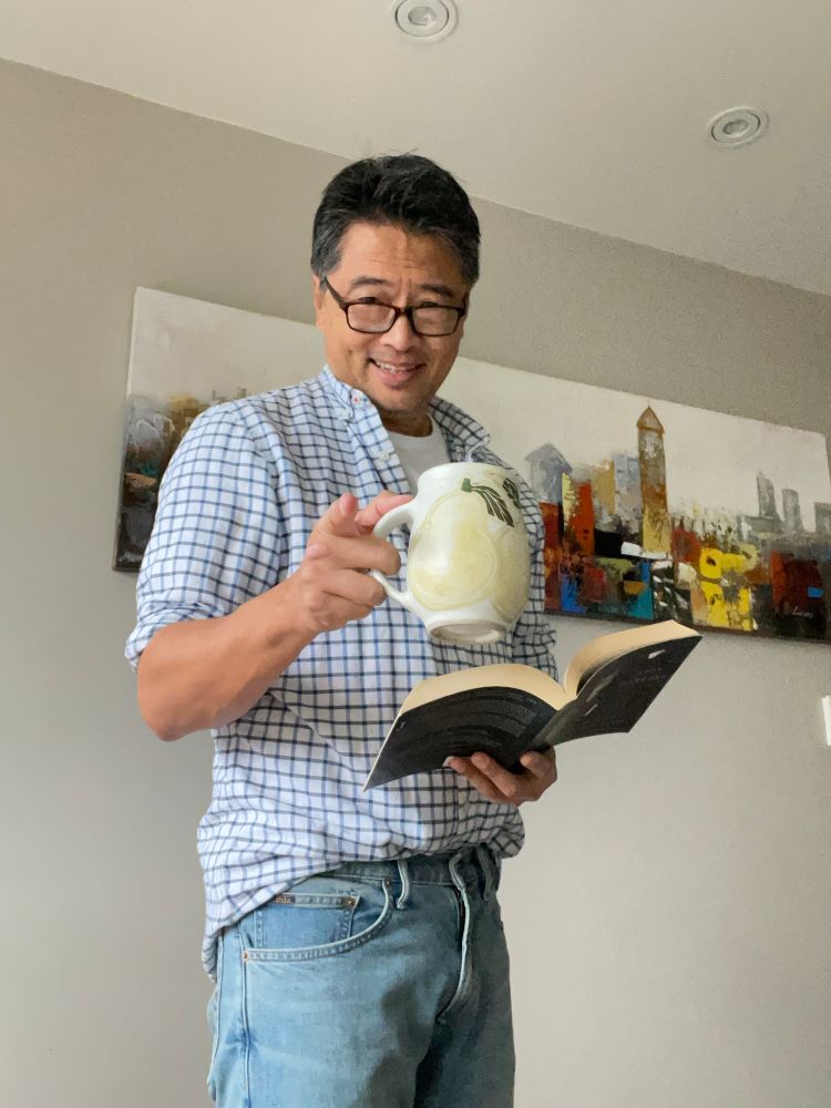 A middle aged Korean man kind of dressed as Jinu from Kpop Demoj Hunters if he was retired. He’s holding a book and cup of coffee. 