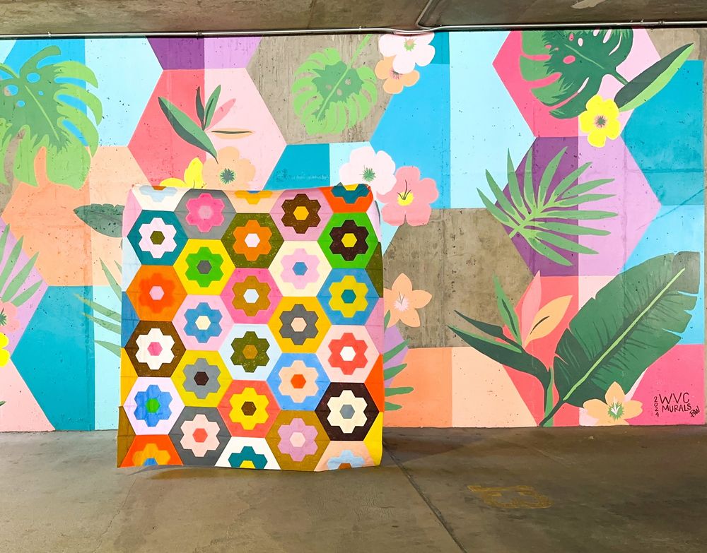 A colorful quilt with EPP hexie flowers on top of large hexagons. It is being held up in front of a mural with colorful hexagons and flowers painted on it