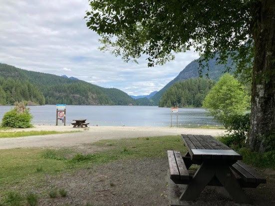 Buntzen Lake, with tree-covered hills and mountains in the background. Bushes and a picnic bench between two signs line the lake shore, and another picnic bench sits under a tree in the foreground.