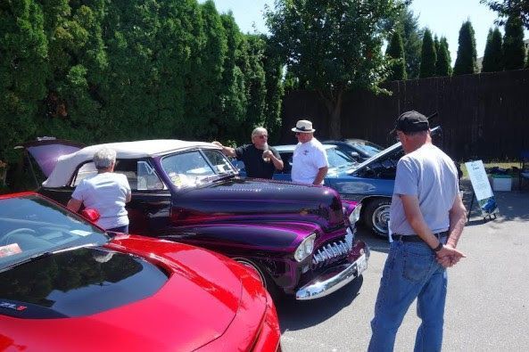 People standing around a purple classic car with its trunk open, parked between a red sports car to its left and a blue classic car with its hood open to its right.