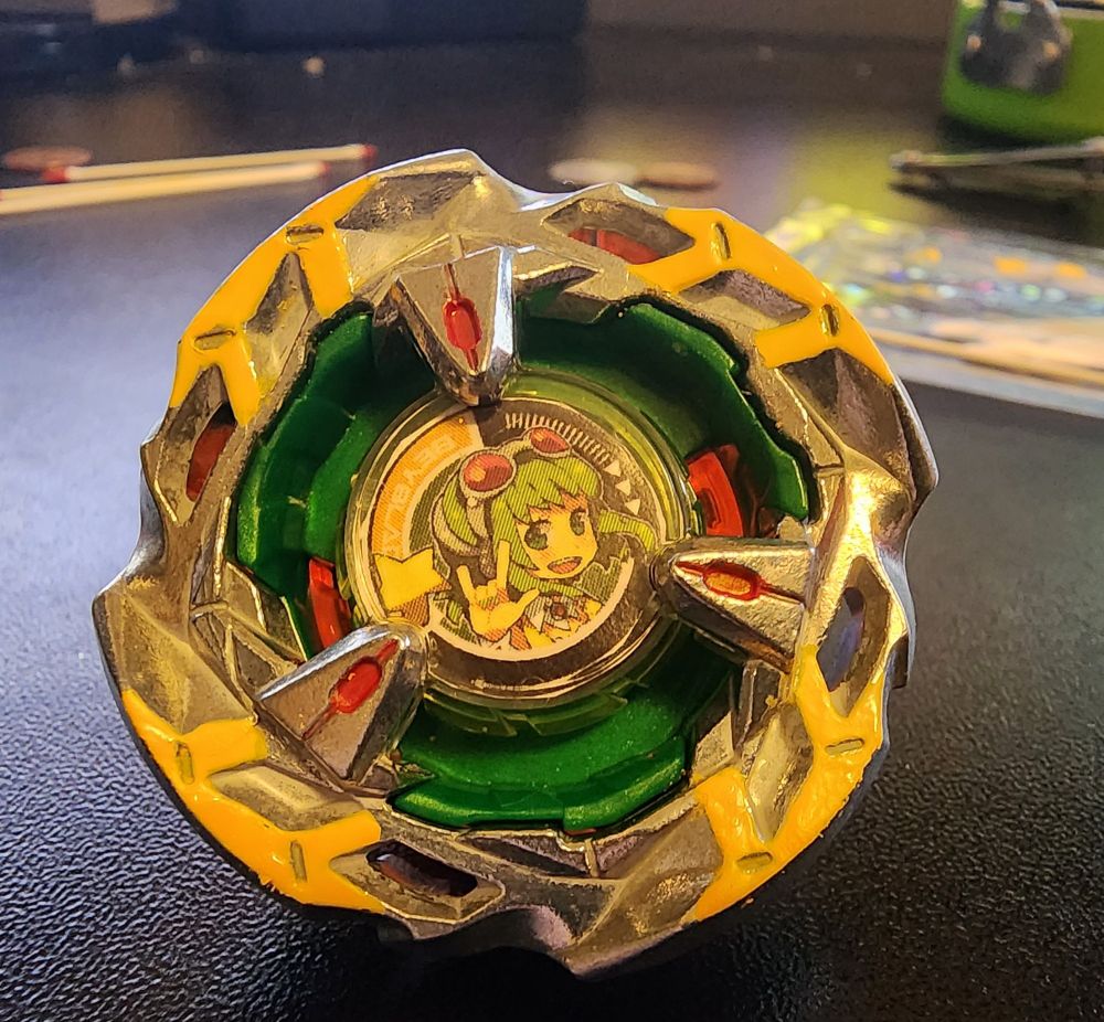 Hells Scythe from the BX-14 random booster, painted yellow on the wings, with red highlights on the prongs. A GUMI Megpoid gearchip sits under the protector. The drawing is attributed to https://www.pixiv.net/users/29325 on danbooru.