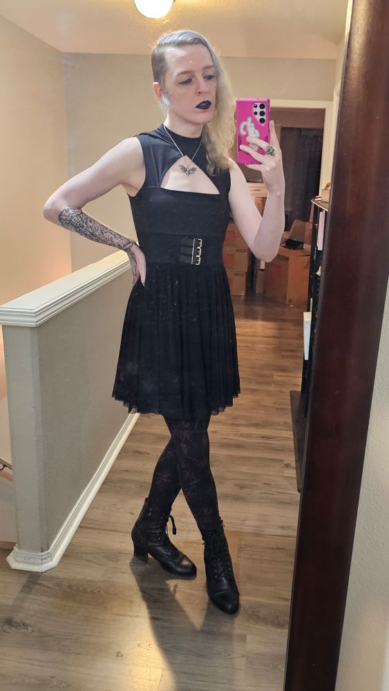 A transfemme in a black dress and black boots, with patterned tights.