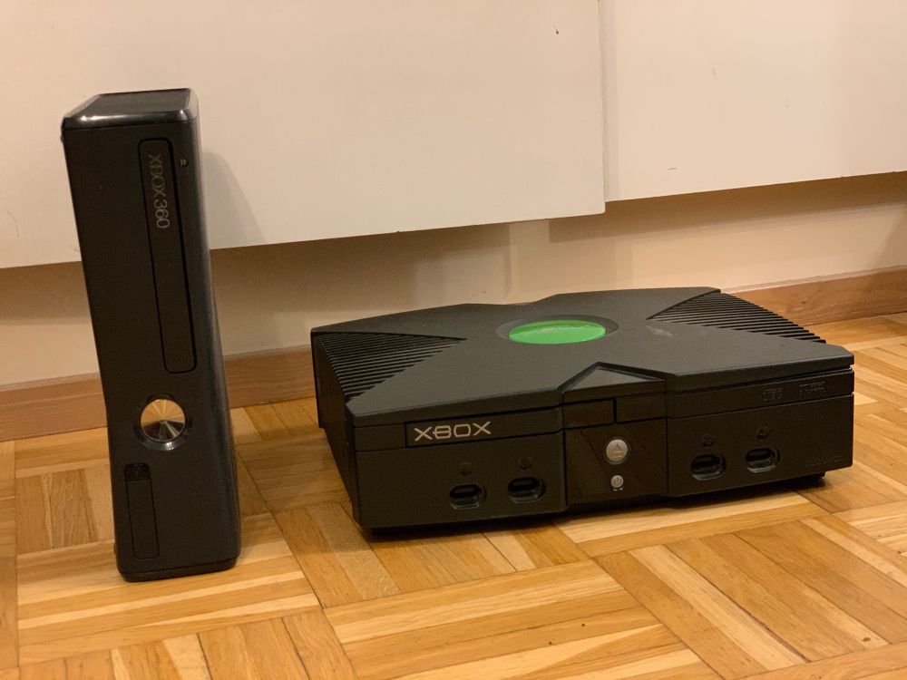 A black XBOX 360 S stood up vertically and a regular black XBOX laying horizontally in its intended orientation in a well lit room on a wooden floor. There is no loss in this picture.