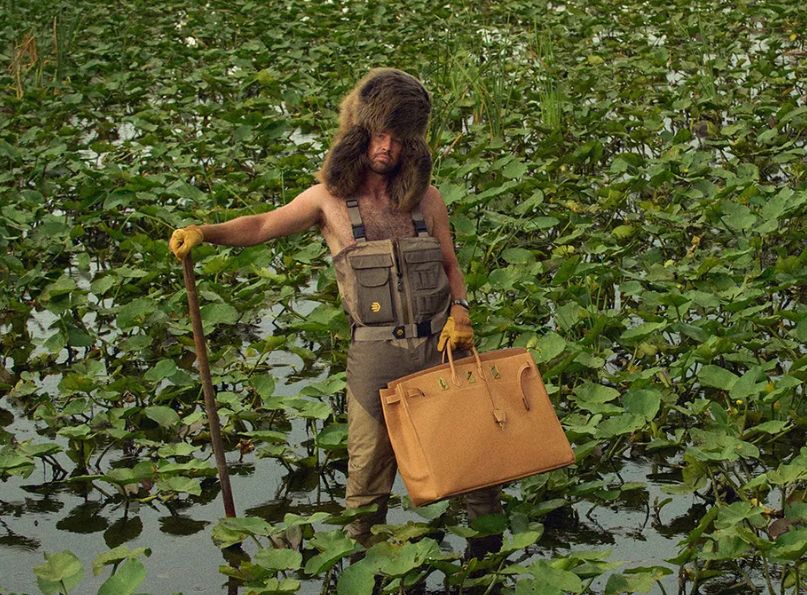 Image of Travis Kelce standing in a Lilypad-filled bog of some sort. He's wearing fishing wader bibs, one of those gigantic fur caps with ear flaps that are resting on his almost-as-hairy chest. He's got a long pole in one hand, and is daintily holding an Hermes Birkin bag that Taylor Swift could literally fit in. And somehow, it all works. God is mysterious, I guess. 