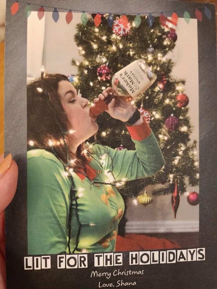 I am in an elf costume pretending to drink directly from a bottle of Maker’s Mark. There are Christmas lights wrapped around me as I sit in front of a decorated Christmas tree. The card says “lit for the holidays”