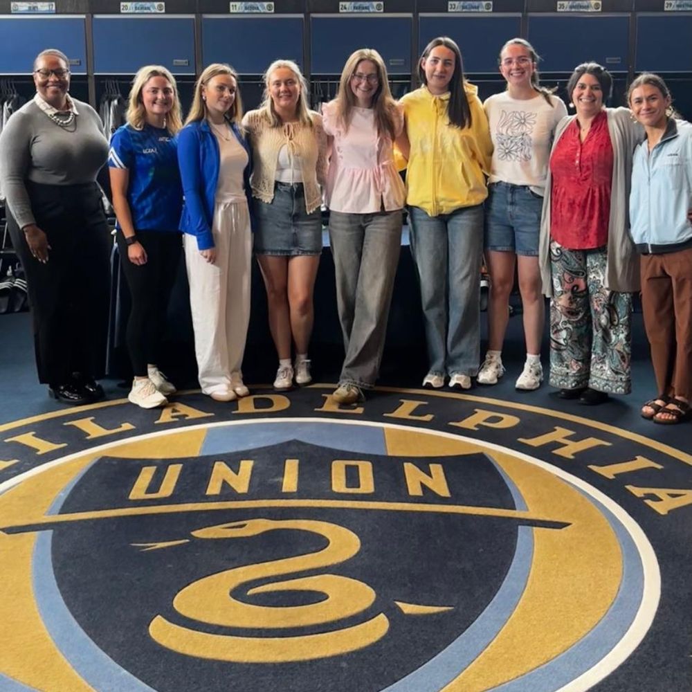 A group of ten adults, all women, stand in a room with a large circular rug featuring the Philadelphia Union soccer club logo. Lockers are visible in the background.