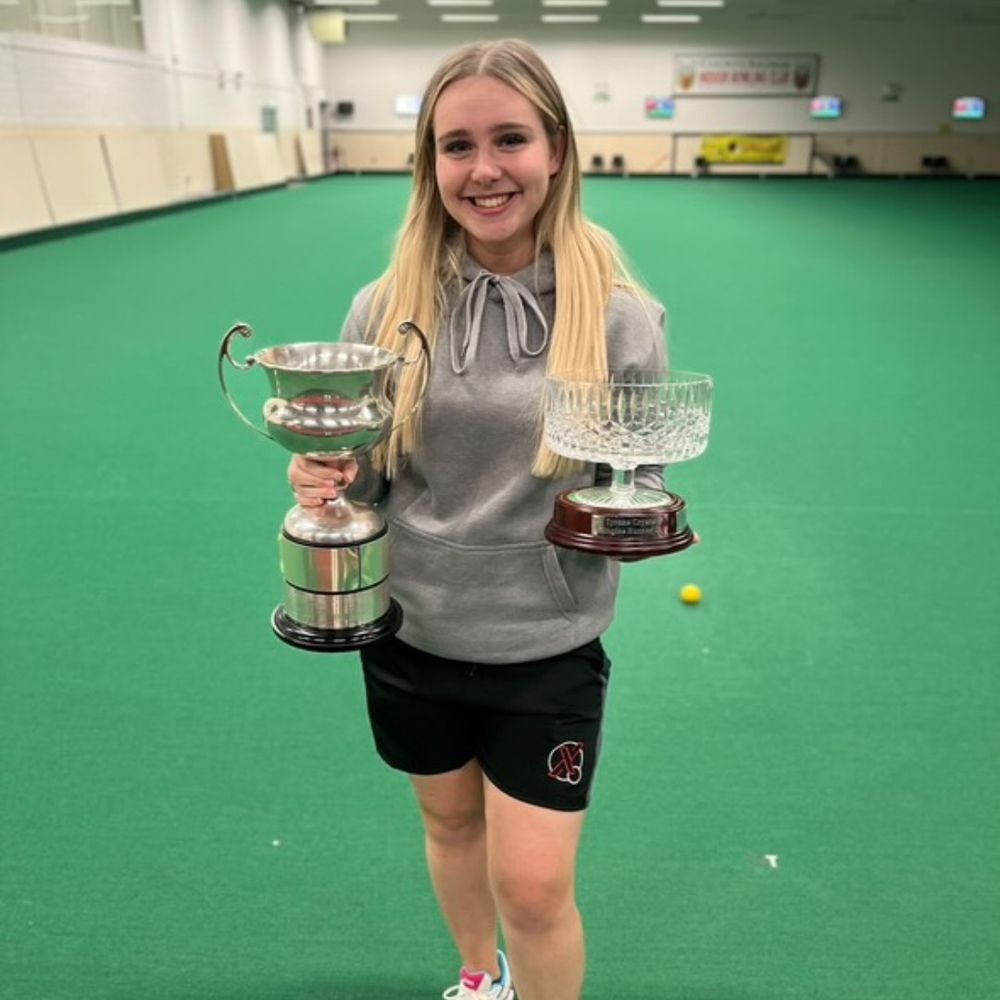 A young woman with long blonde hair stands in the center of an indoor bowling green, smiling and holding two trophies. The trophy in her left hand is a large silver cup, and the one in her right is a crystal bowl. She is wearing a grey hoodie and black athletic shorts, with a green bowling surface extending far behind her.