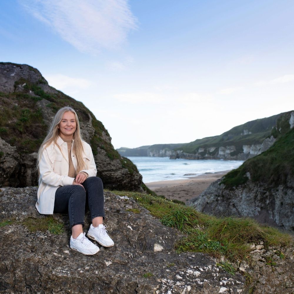Student with long blonde hair wearing a white jacket and sneakers sitting on a rocky cliff overlooking a sandy beach and ocean waves.